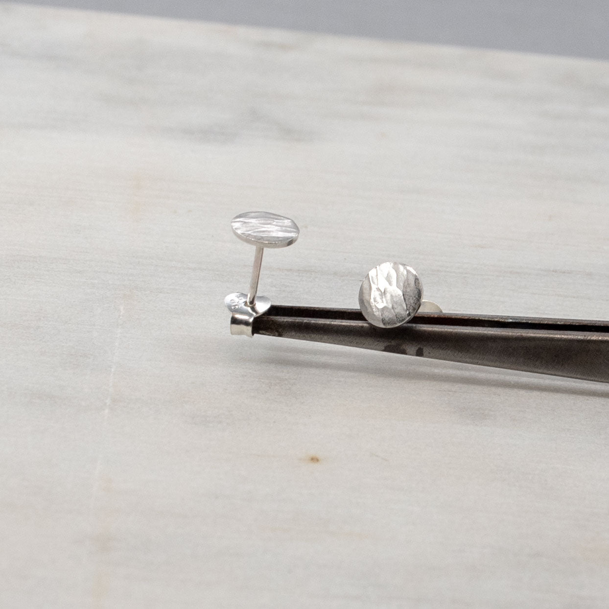 Image en gros plan de deux boucles d'oreilles en argent sterling posées sur une paire de pincettes. Les deux boucles d'oreilles présentent une finition texturée et martelée. L'arrière-plan est d'une couleur douce et neutre, mettant en valeur les boucles d'oreilles.