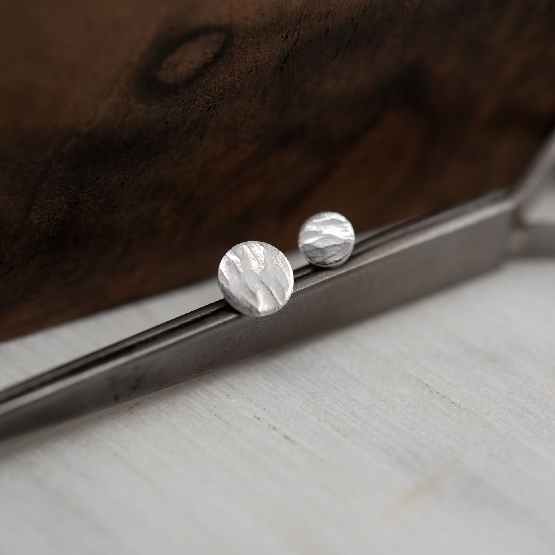 Boucles d'oreilles asymétriques en argent martelé posées sur un outil métallique, avec un fond en bois sombre. Design artisanal minimaliste, idéal pour un style naturel et quotidien.