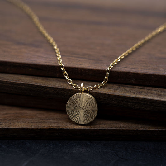 Vue de face collier disque en vermeil doré sur fond bois. Pendentif minimaliste intemporel et élégant gravé à la main. 