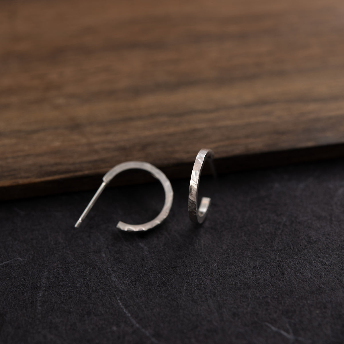 Paire de créoles ouvertes en argent texturé, posée sur une surface sombre texturée, avec un arrière-plan en bois.