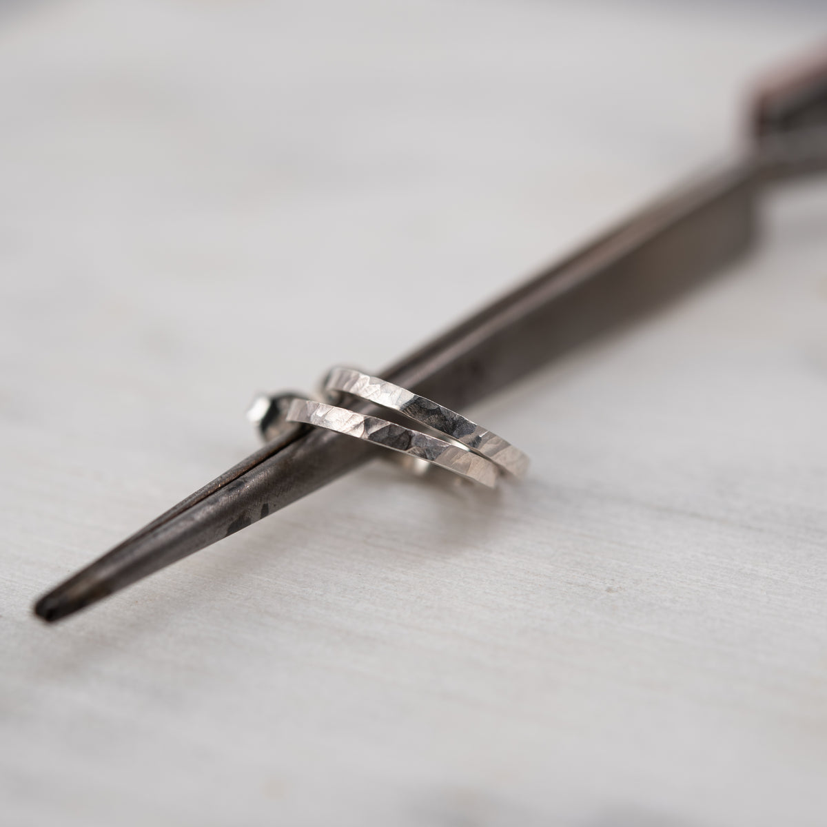 Two hammered silver square wire hoops resting on a dark metal tool against a light gray textured background.