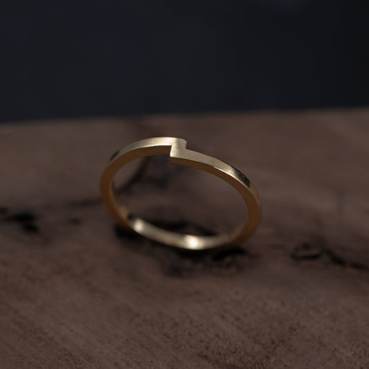 A minimalist gold ring with a unique open-ended, stepped design, resting on a dark brown textured wooden surface against a dark background.