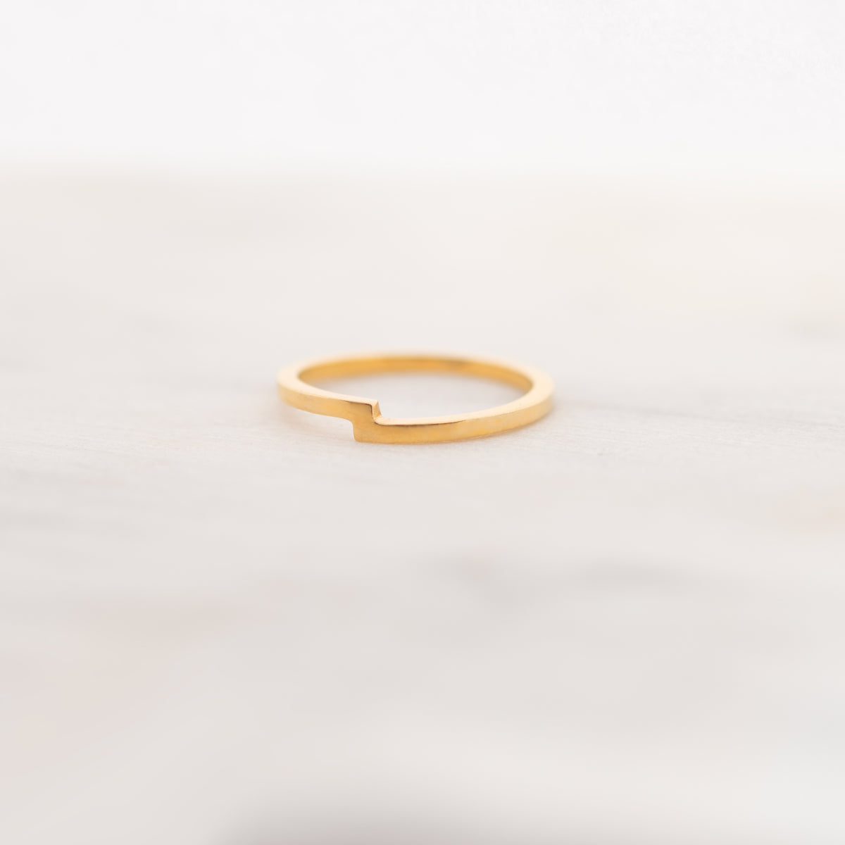 A minimalist gold ring with a unique open-ended, stepped design, resting on a light marble surface against a soft white background.