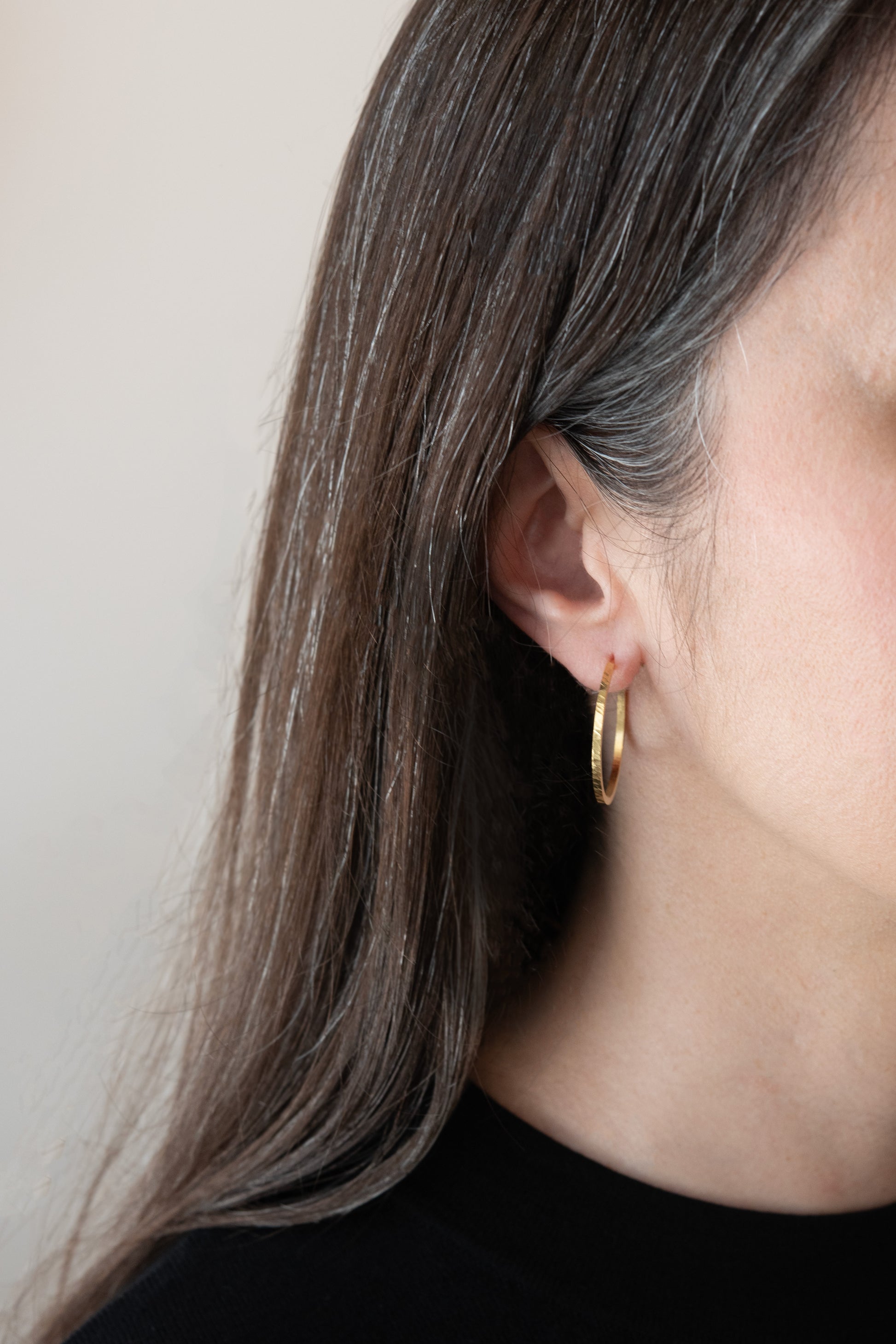 Close-up of a person wearing a gold hoop earring against a neutral background -light-catching handmade jewelry