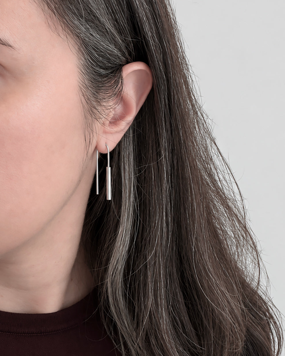 Gros plan sur l’oreille d’une femme portant une paire de longues boucles d’oreilles pendantes minimalistes en argent, avec des éléments cylindriques, sur un fond gris clair.