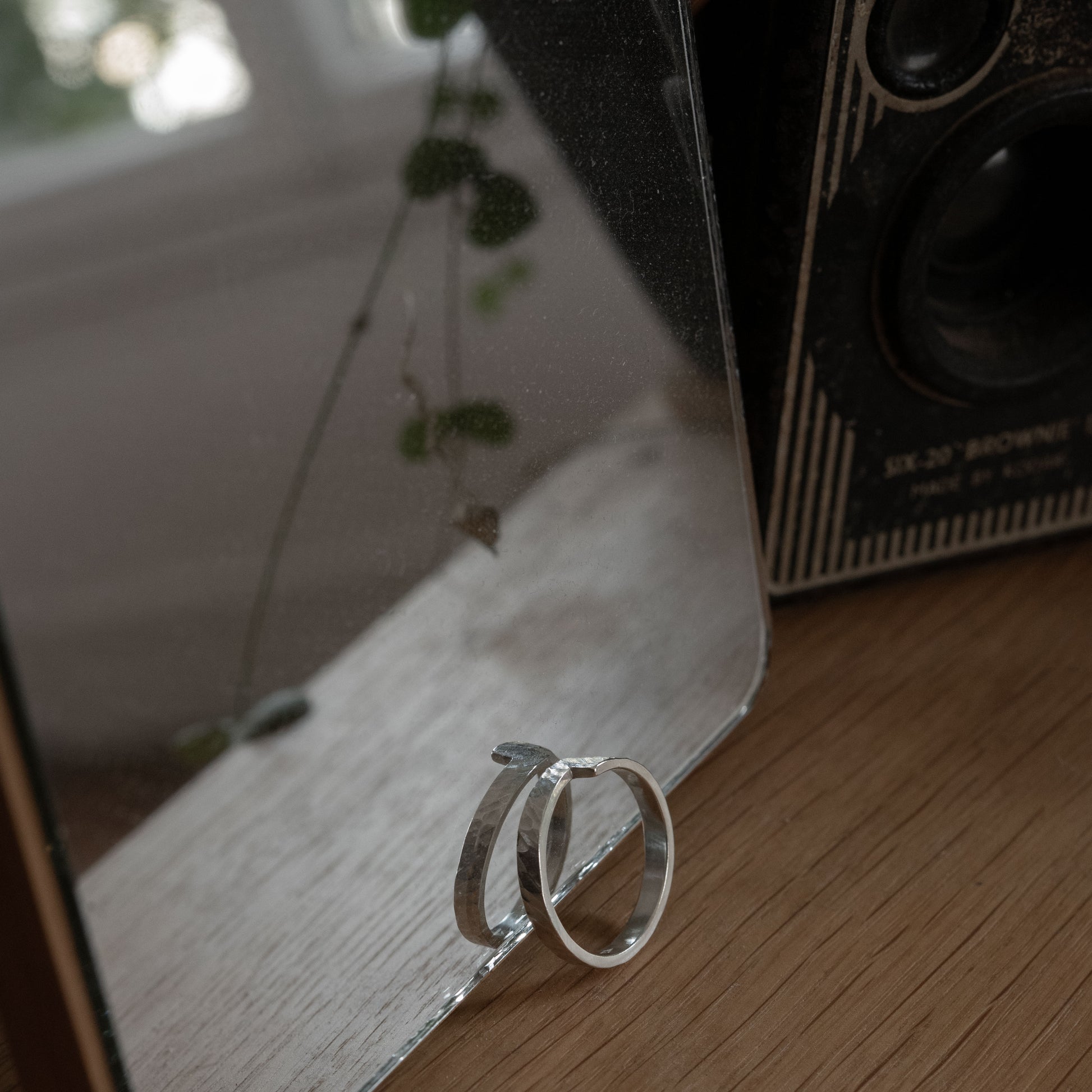 Artistic view of the Hammered Crossed Band Ring in sterling silver, displayed on a wooden surface next to a reflective mirror. The ring’s unique design is highlighted by the mirror’s reflection, with greenery visible in the background, adding a natural touch to the composition.