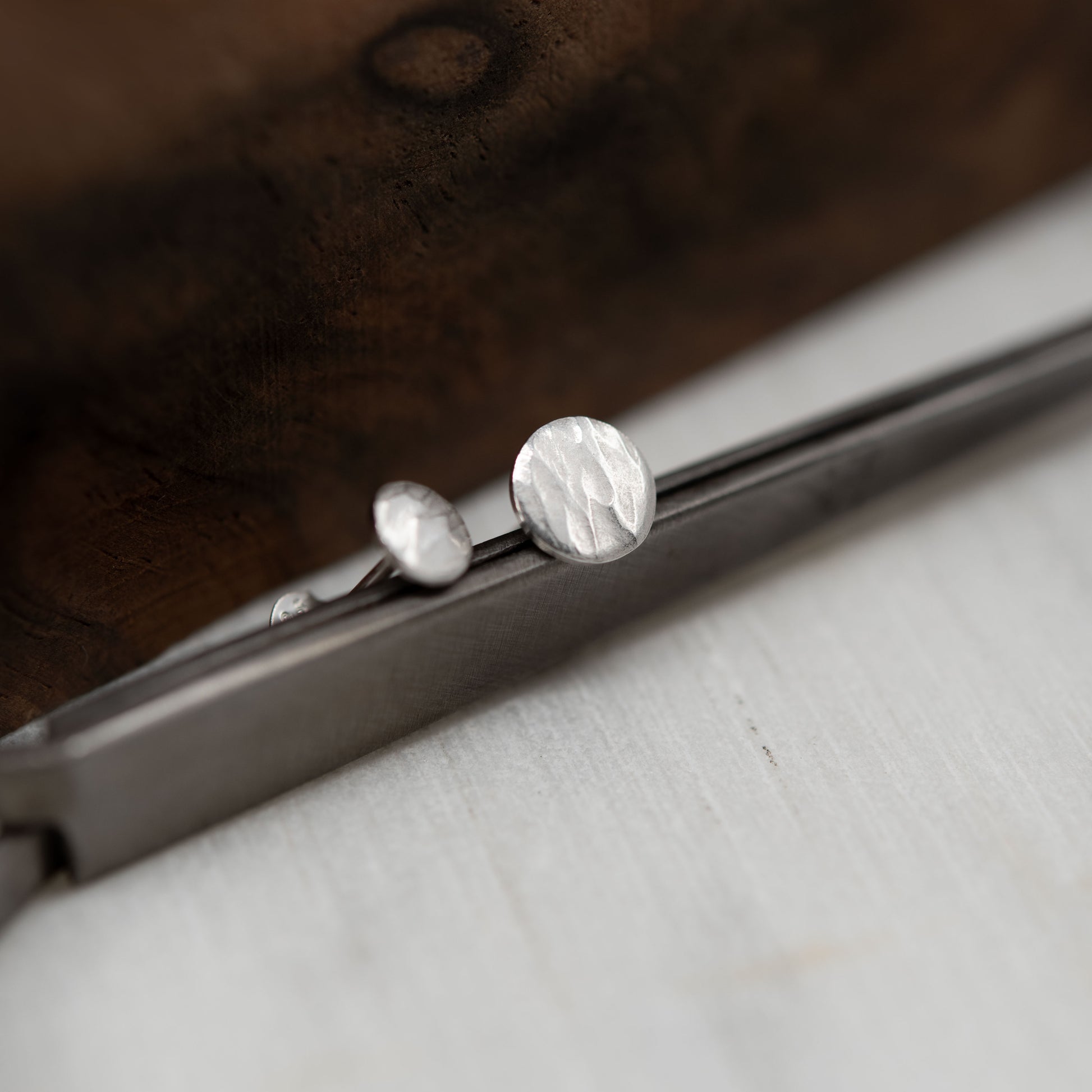 Close-up of mismatched hammered silver stud earrings displayed on a metal tool with a wooden background. Features a natural, handcrafted texture and minimalist design.