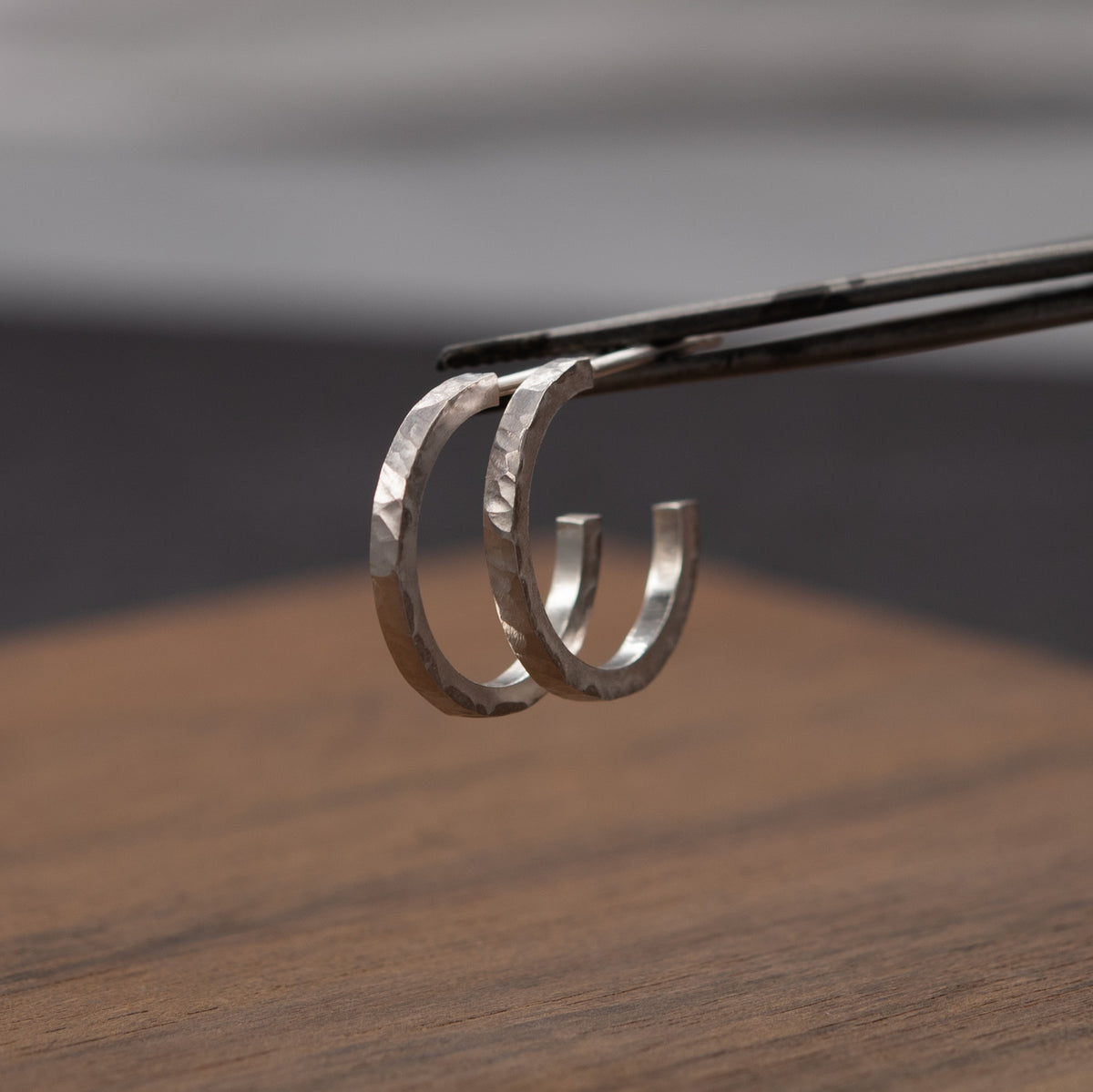 A pair of hammered silver hoop earrings handcrafted in Paris, held by black tweezers over a blurred wooden surface and gray background.