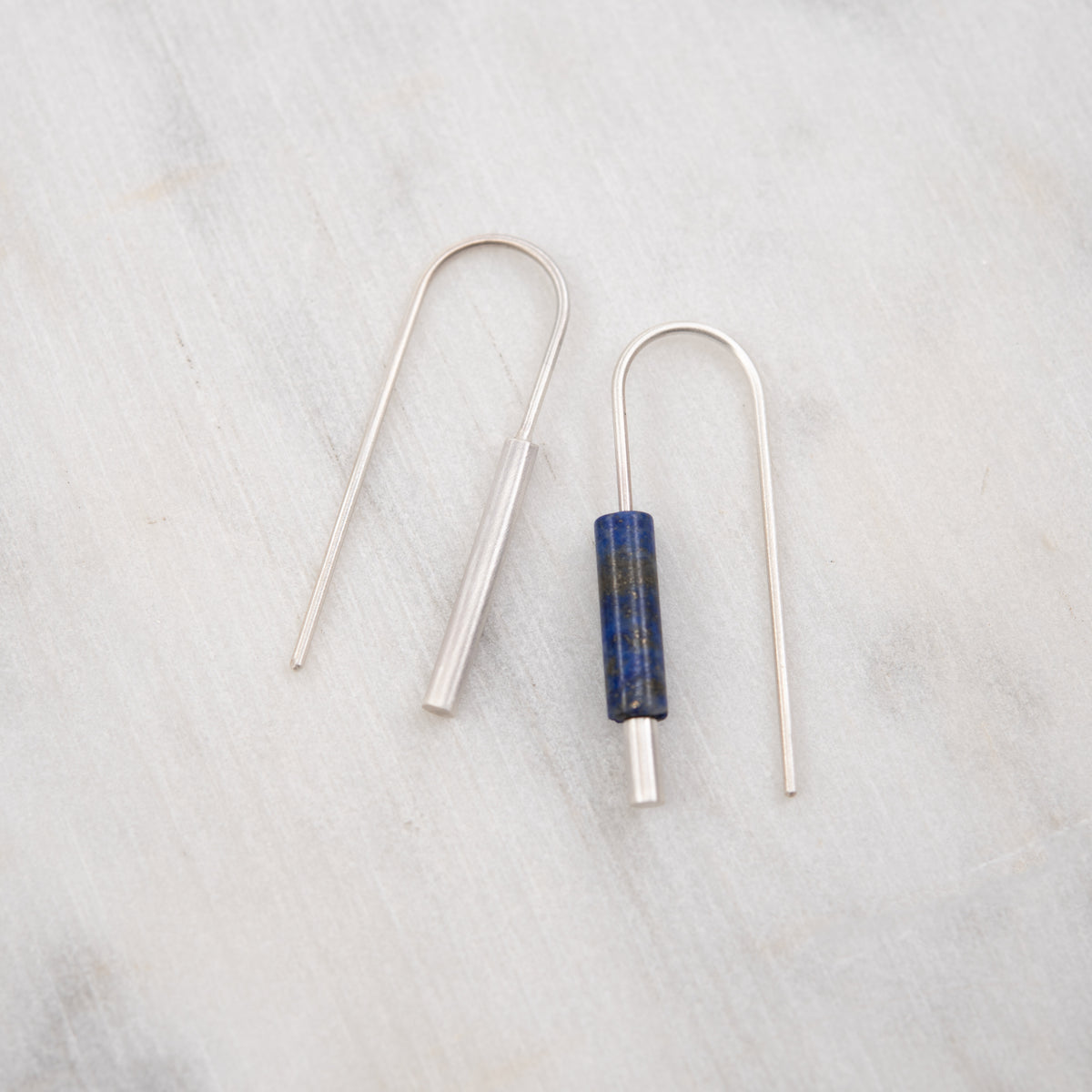 A pair of minimalist silver-toned drop earrings on a light gray marble surface. One earring is plain, and the other features a cylindrical blue lapis lazuli stone bead in the center.