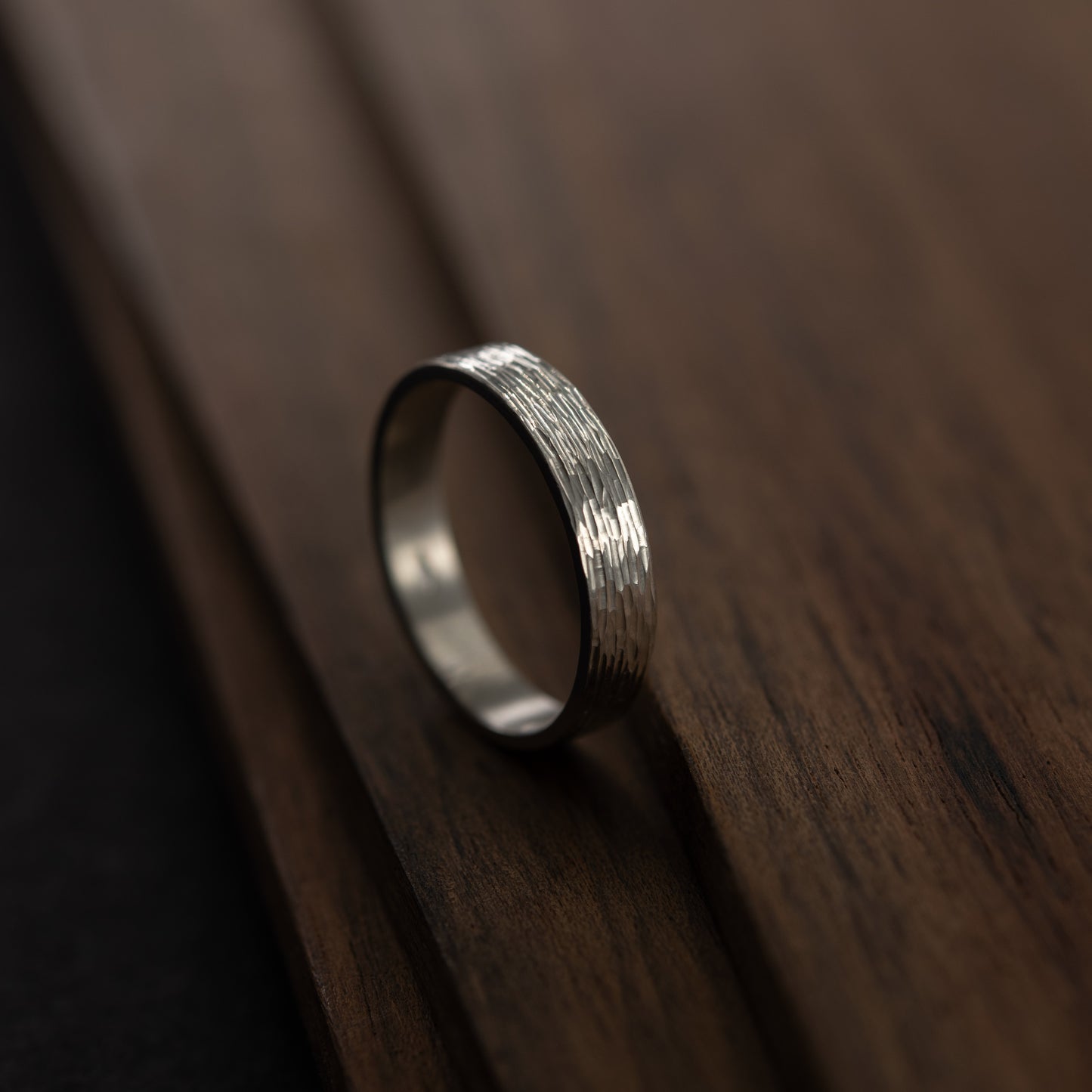 Side view of handmade silver wedding band with hand-engraved texture standing upright on wood, warm minimal setting.