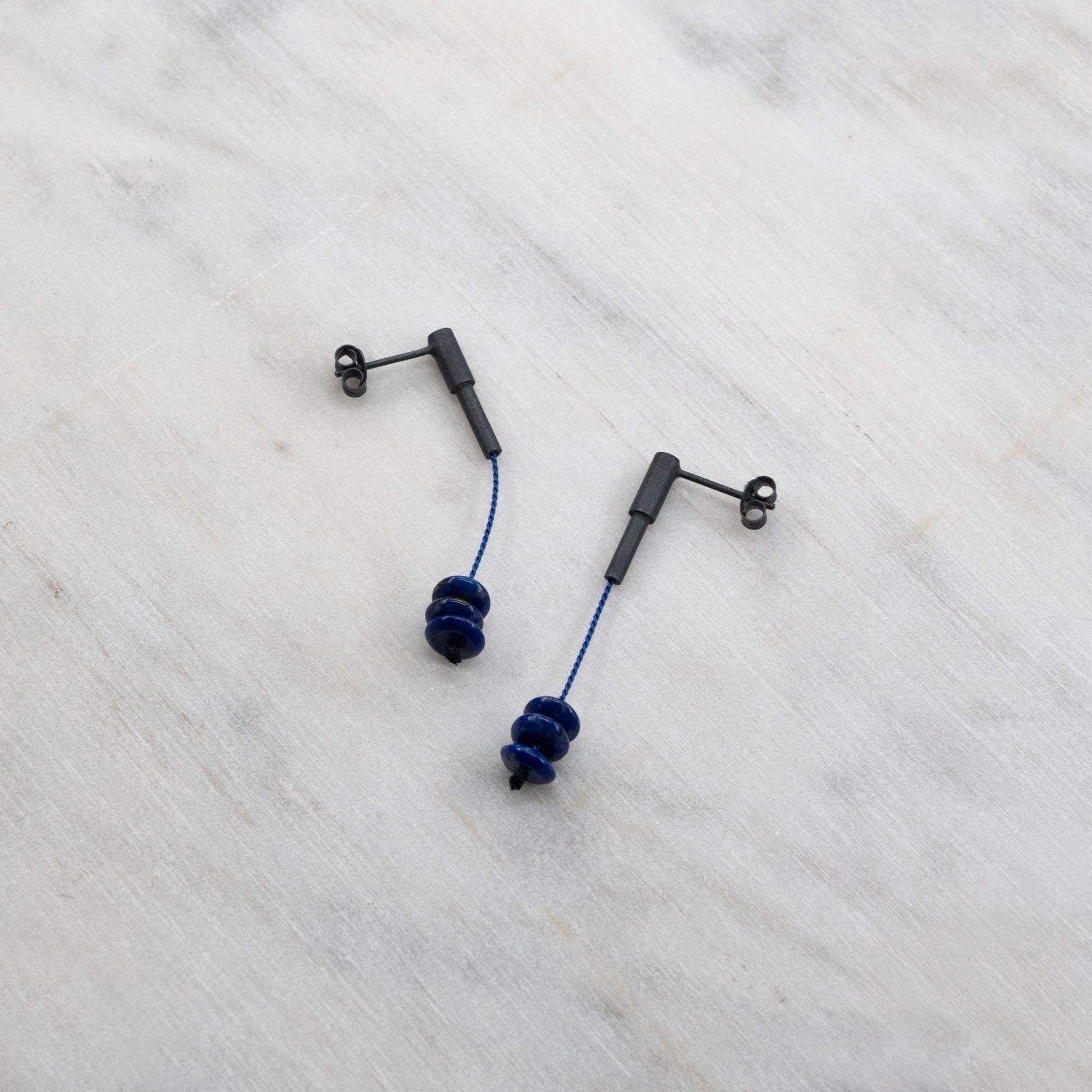 View of lapis lazuli earrings crafted in oxidized silver, featuring blue silk cord, elegantly displayed on a marble surface. The earrings showcase their vibrant color and unique design, adding a touch of sophistication to the composition."