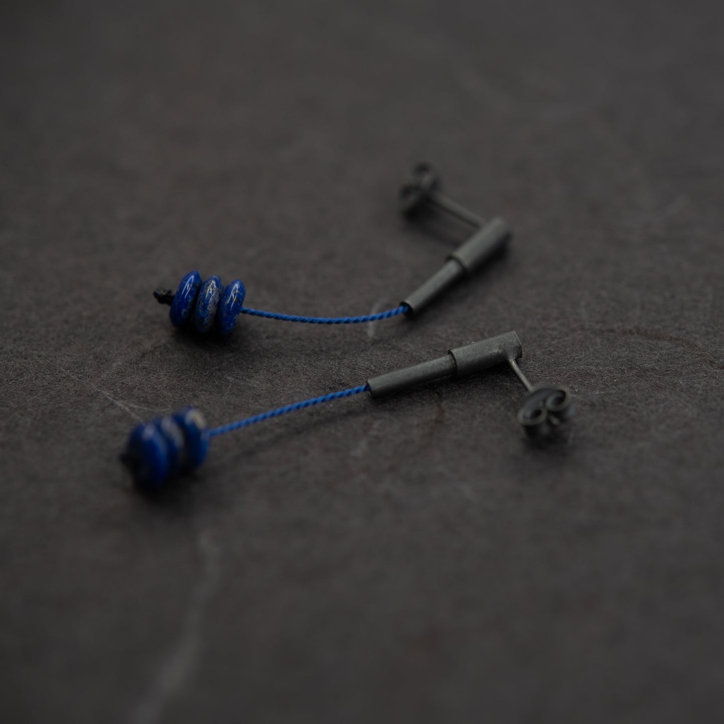 Close-up of lapis lazuli earrings crafted from oxidized silver, featuring blue silk cord, displayed on a dark textured background. The vibrant blue beads are stacked, highlighting their unique design and modern aesthetic.