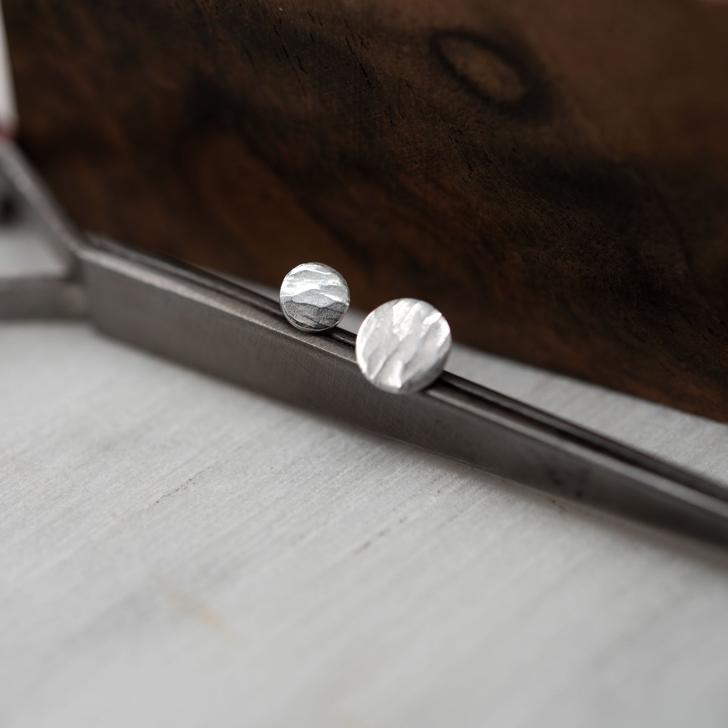 Mismatched hammered silver stud earrings displayed on a metal tool with a wooden background. Features a handcrafted organic texture and minimalist design.