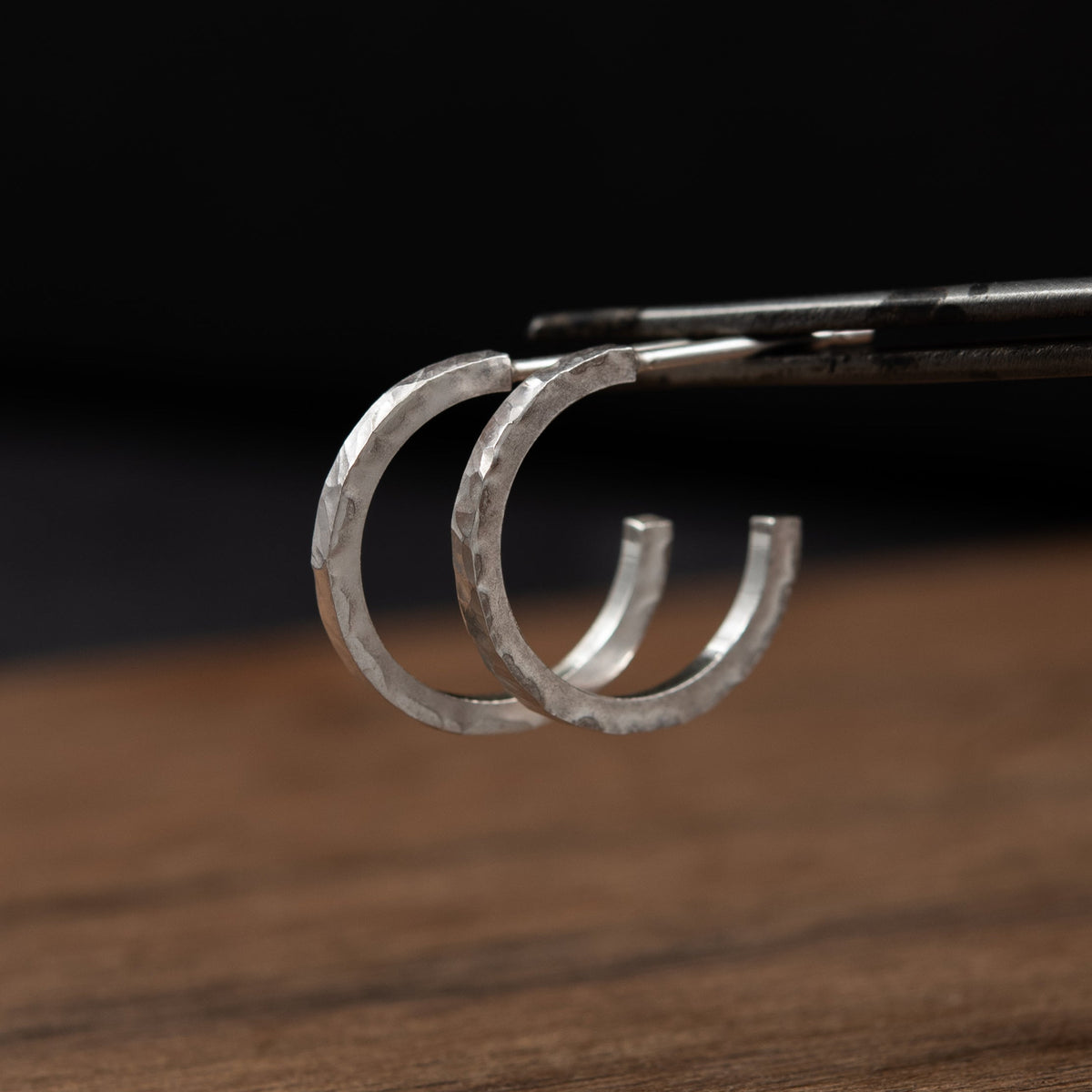 Deux petites créoles en argent massif  texturé martelée, tenues par une pince métallique au-dessus d’une surface en bois avec un arrière-plan sombre et flou.