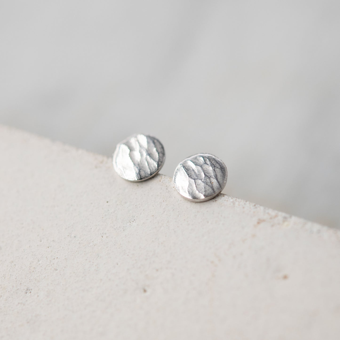 Image en gros plan de deux boucles d'oreilles en argent sterling posées sur un bord de surface claire. Les boucles d'oreilles ont une forme ronde avec une finition martelée qui capte la lumière, mettant en valeur leur texture artisanale. L'arrière-plan est flou et doux, accentuant l'élégance des bijoux.