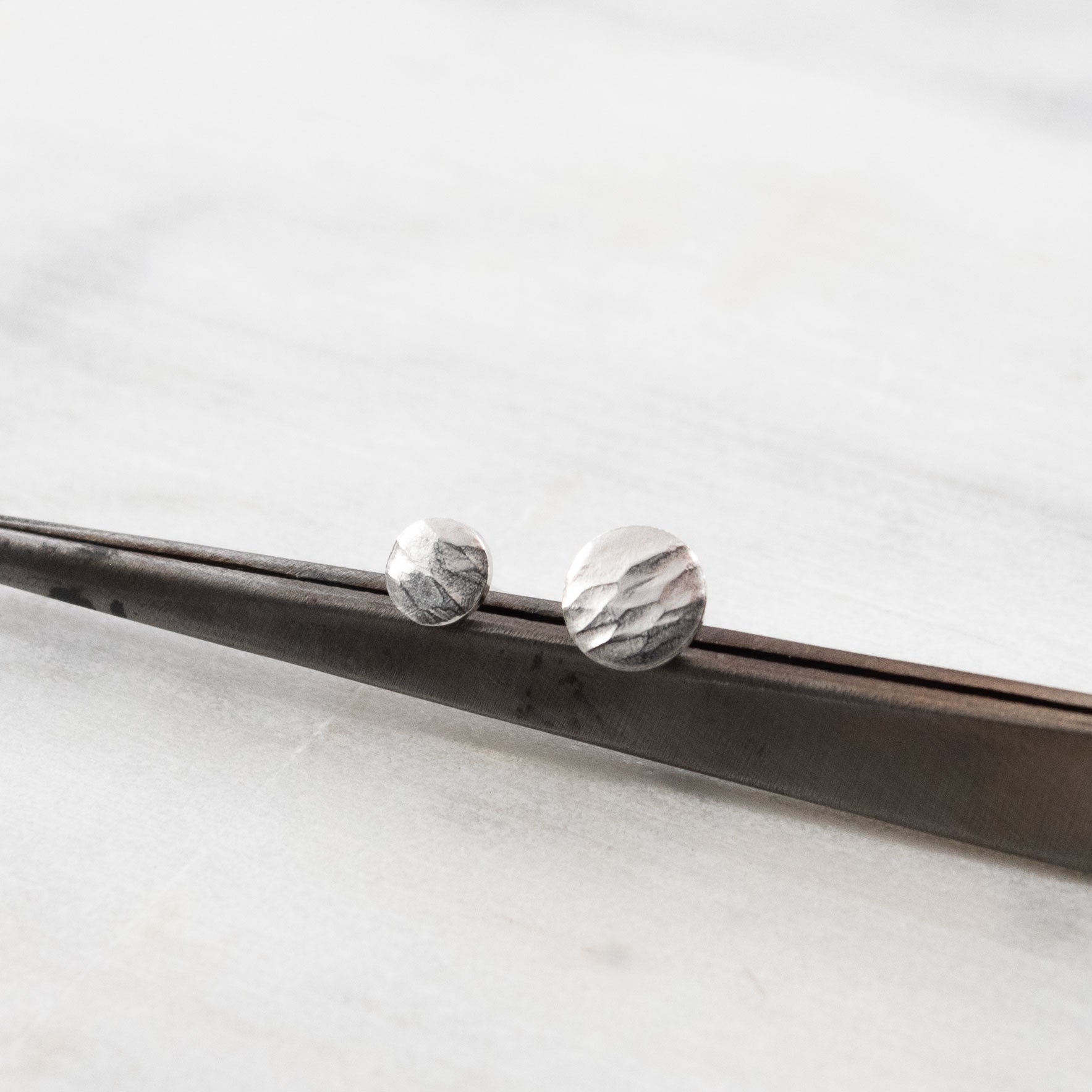 Vue rapprochée de boucles d'oreilles asymétriques en argent martelé, posées sur un outil en métal. Design minimaliste et artisanal avec une texture naturelle et organique.
