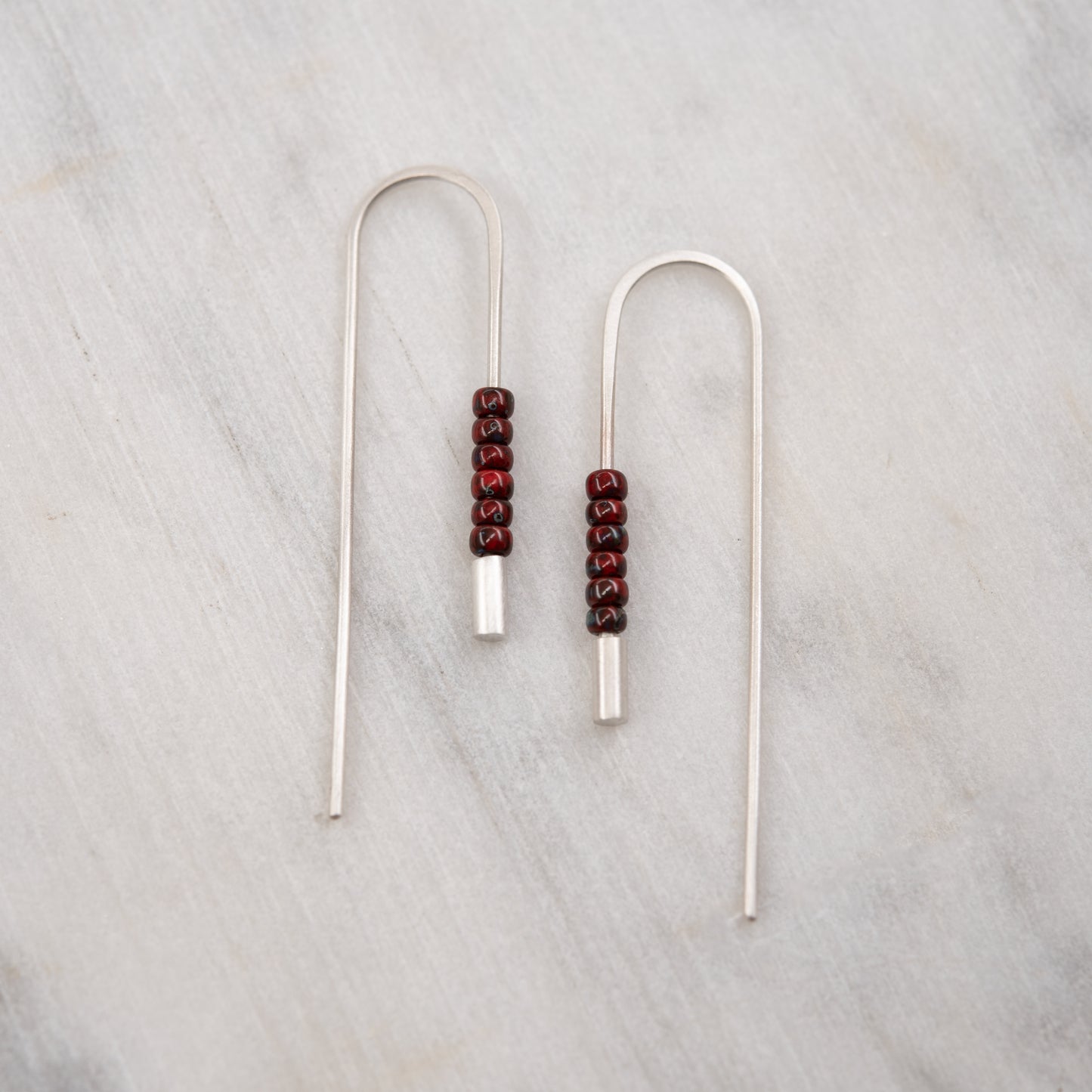 Front view of long brushed sterling silver threader earrings decorated with red Miyuki beads on a light background