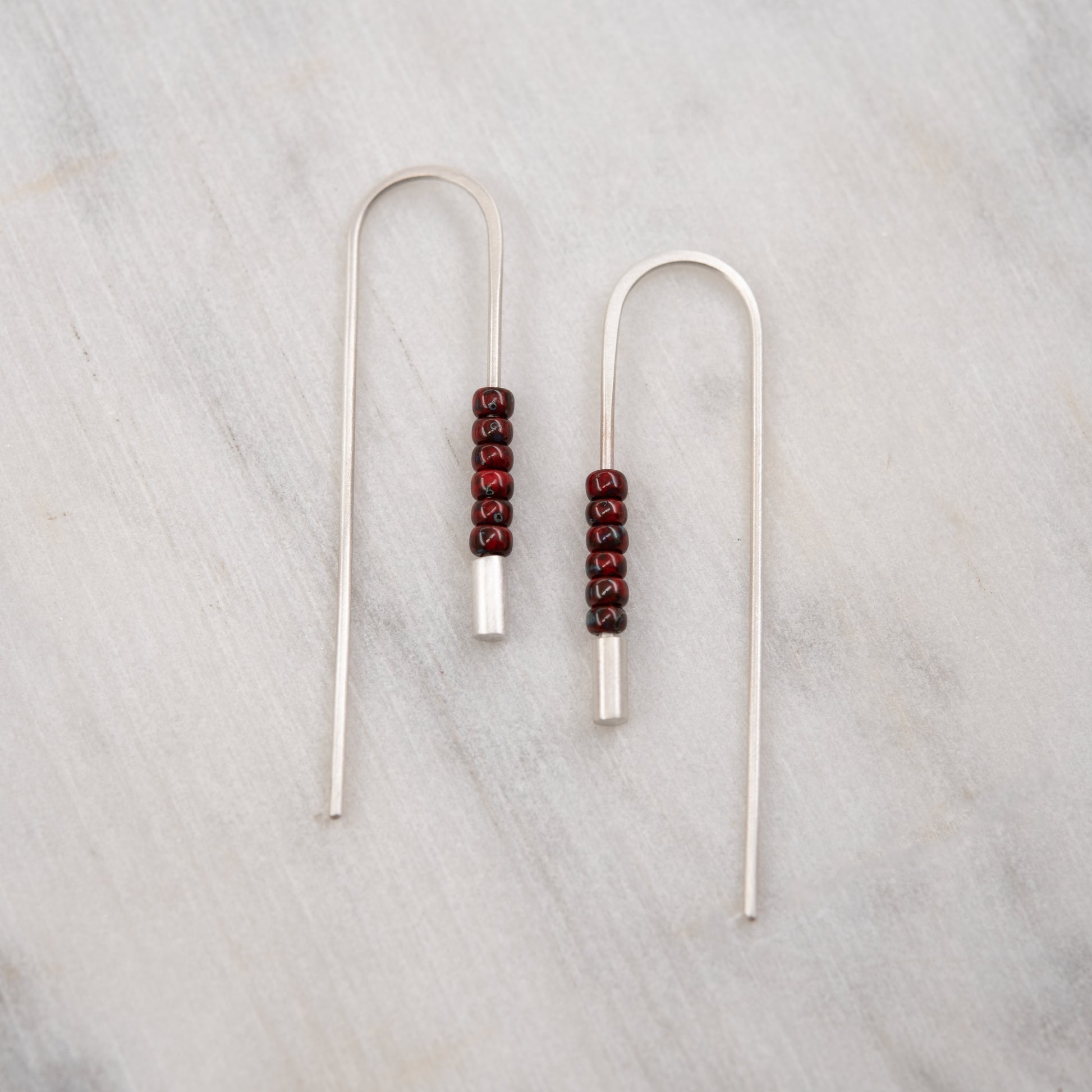 Front view of long brushed sterling silver threader earrings decorated with red Miyuki beads on a light background