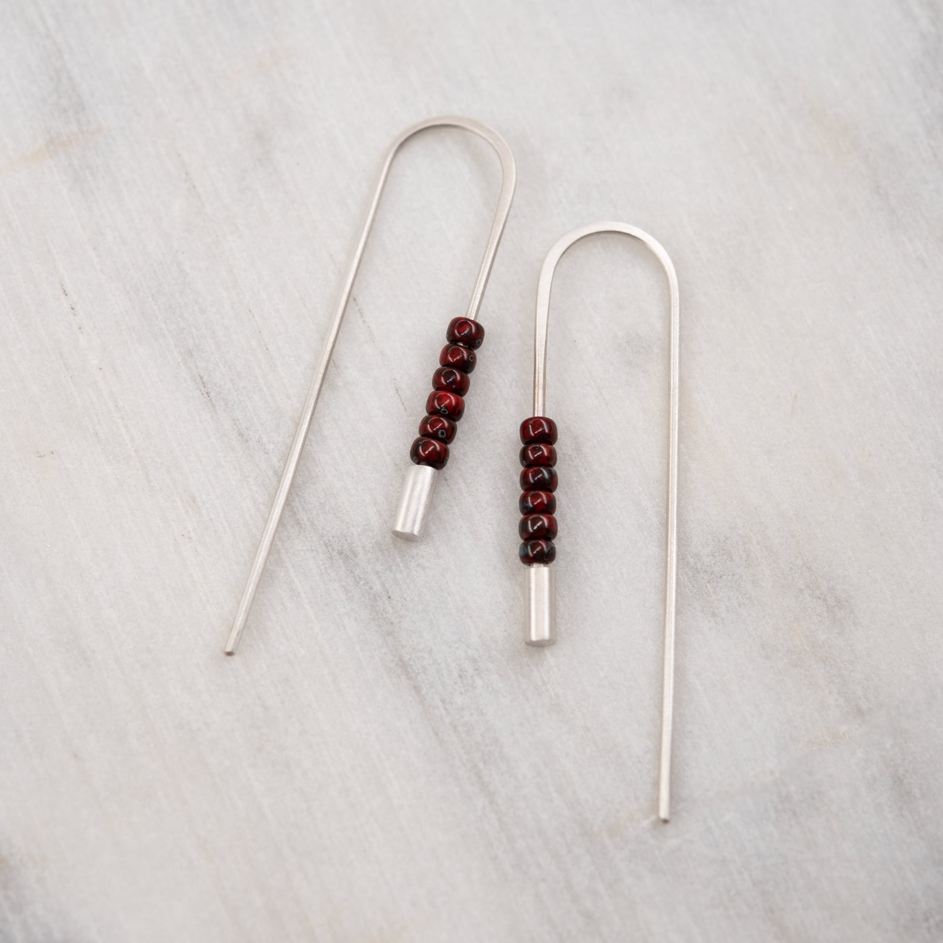 Pair of brushed sterling silver threader earrings with red Miyuki beads on a pale marbre background
