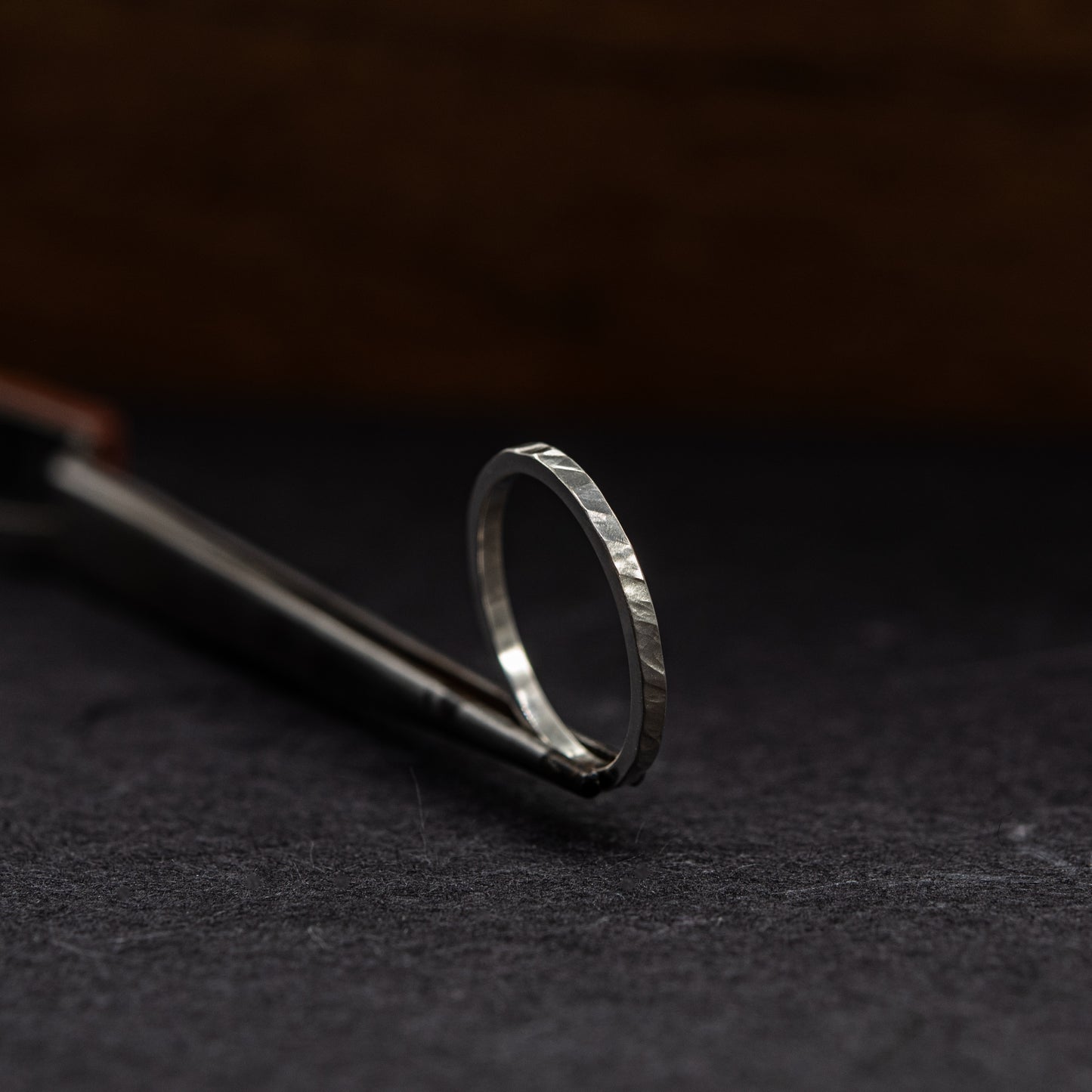 A silver ring with a textured, hammered finish resting on a dark surface next to a metal tool.