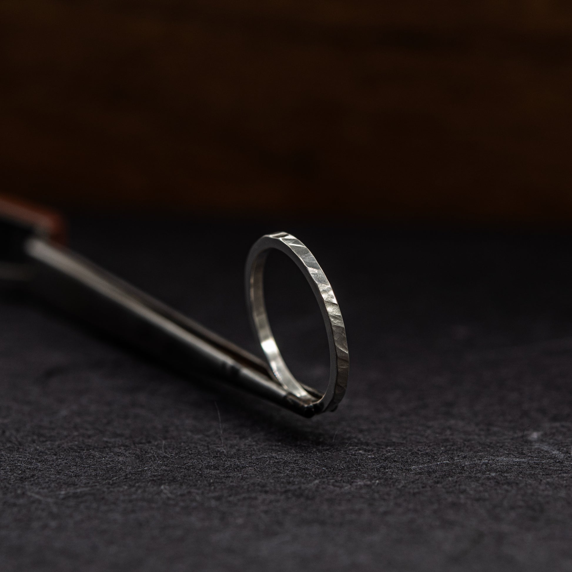 A silver ring with a textured, hammered finish resting on a dark surface next to a metal tool.
