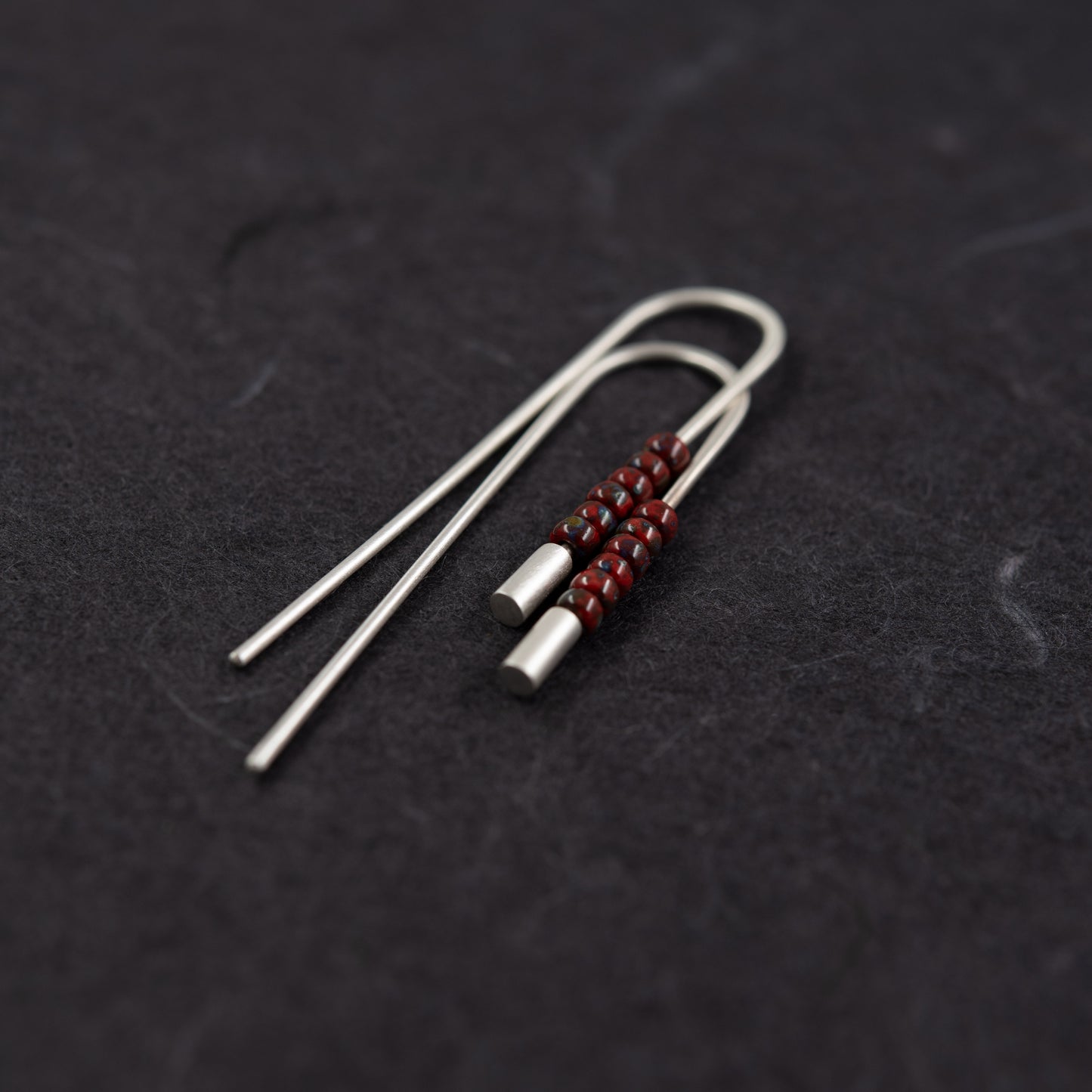 Close-up of handcrafted sterling silver threader earrings with red Miyuki beads on a dark background