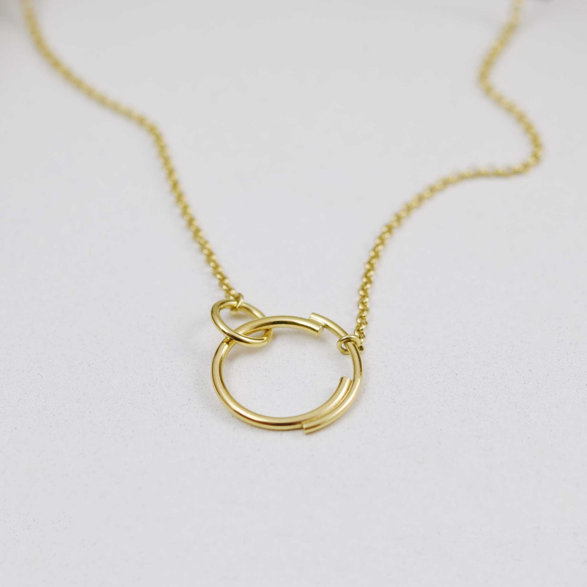 A close up of a stylish interlocking circle necklace  in gold that features a small circle clasping onto a larger circle that is made up of two separate arcs for a truly eclectic design.