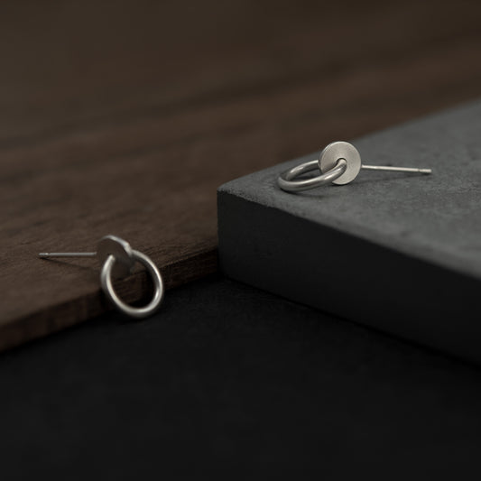Close up of a pair of silver matte studs designed with an interlocked dot and circle  The earrings measure 13 millimeters in length.