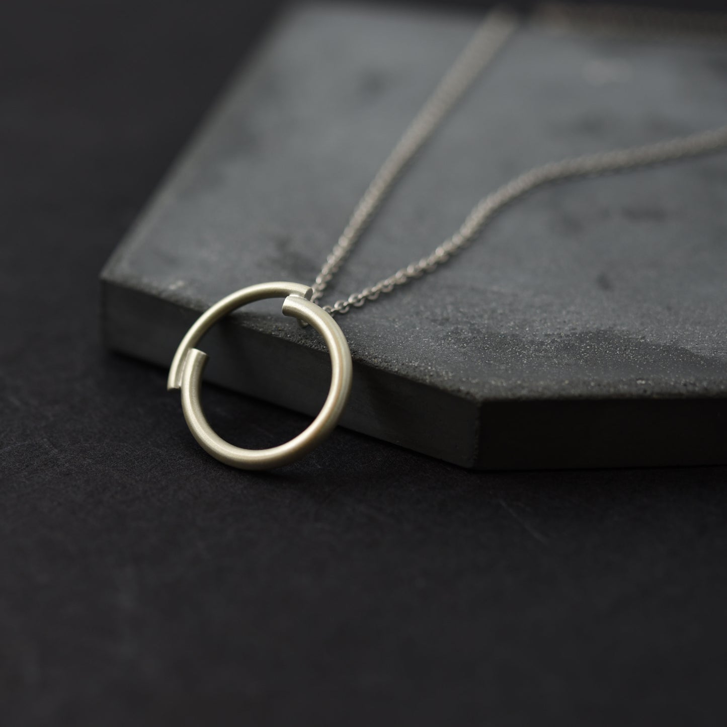A close up of a silver circle pendant necklace of 22 millimetres diameter in a brushed matte finish in a dark blue background  