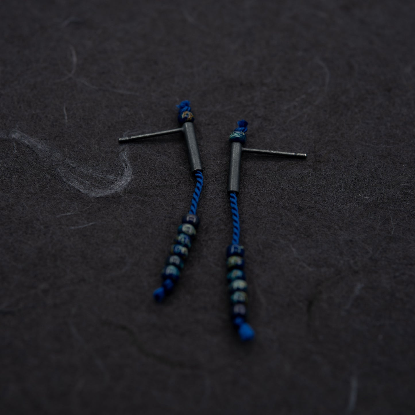 Image showing a pair of 38 mm length pendant earrings handmade in oxidized black sterling silver tube with a blue silk cord threaded through the center for hanging Picasso blue Miyuki beads. Designed and made by hand in Paris by A g J c 