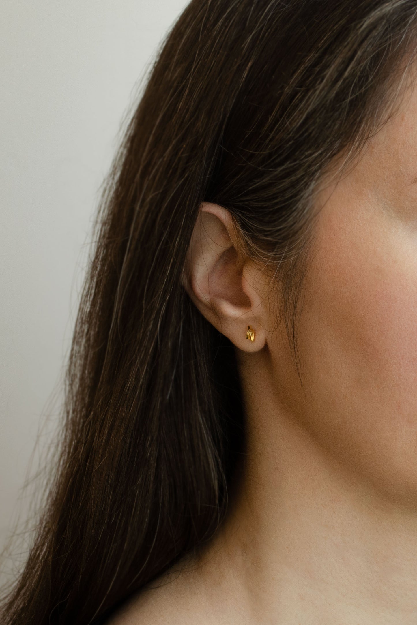 A half face of a model with long brown hair wearing a Tiny loop studs earrings crafted by hand from a 1,5 mm square wire of sterling silver and then shiny finished and gold plated. Length: 5 Millimeters; Width: 7 Millimeters