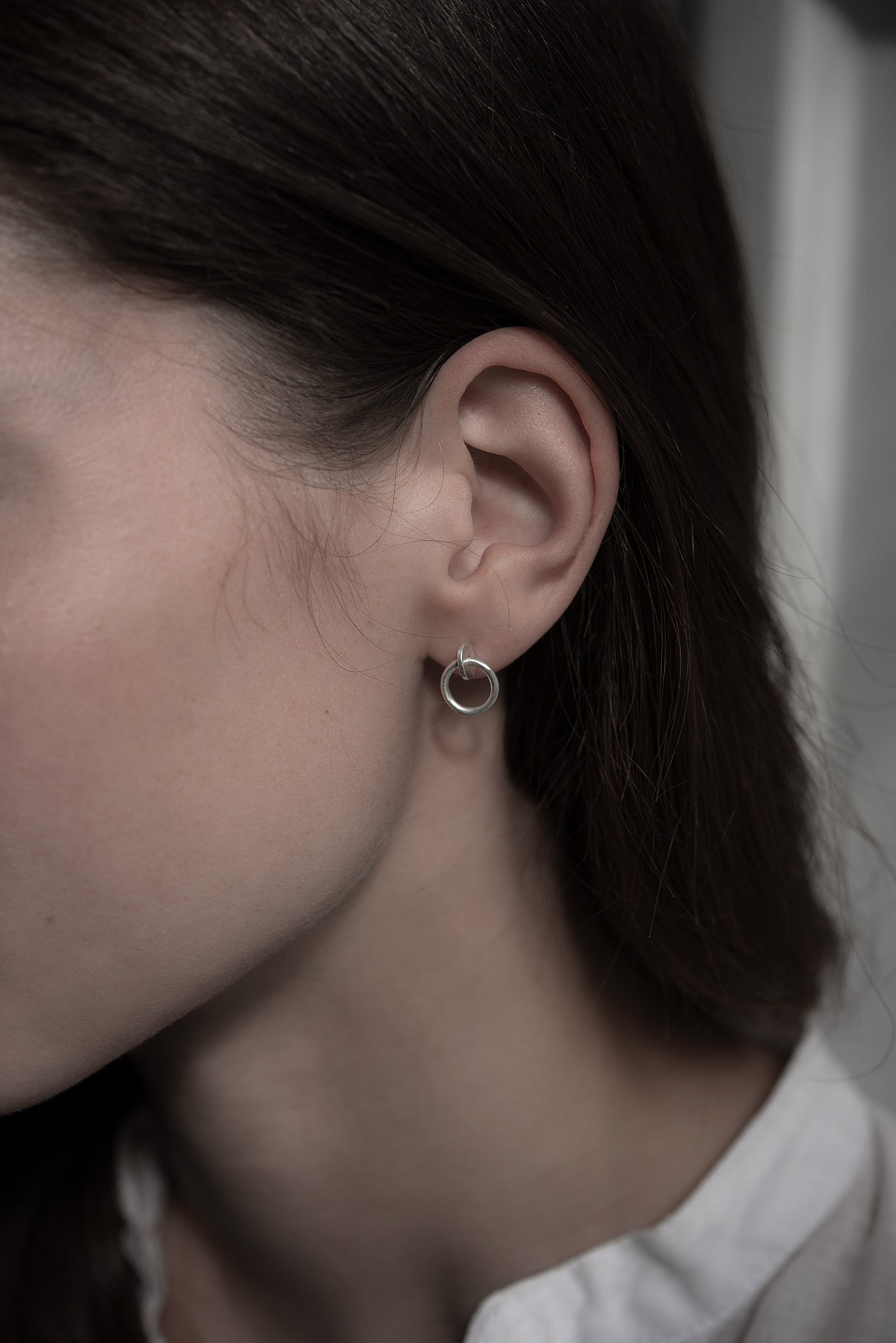 Close up of a A woman's face wearing a pair of matte silver studs designed with an interlocked dot and circle  The earrings measure 13 millimeters in length.